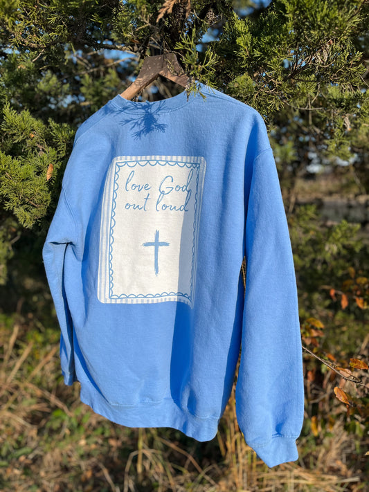 Blue sweatshirt with a white patch and cross design hanging on a tree branch.