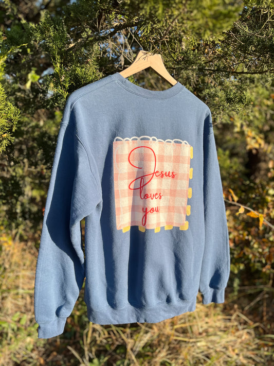 Blue sweatshirt with 'Jesus loves you' design on a hanger against a natural background